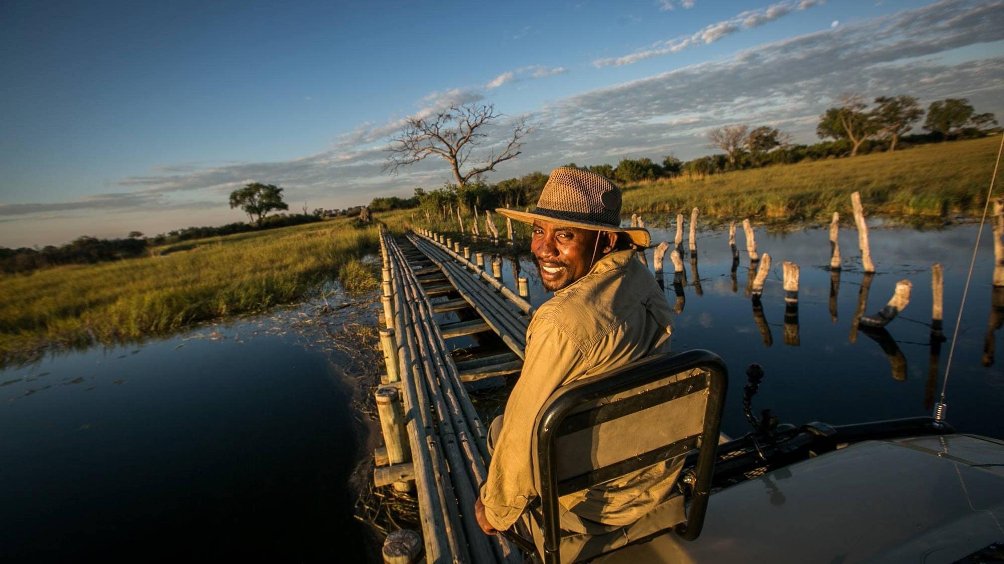 Safari jeep rijdt over een houten brug in de Okavango delta met een spotter die op een stoeltje voor op de motorkap zit